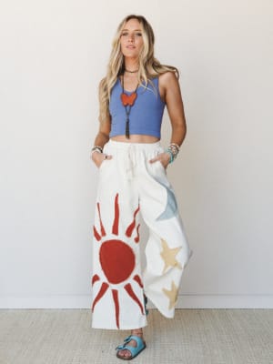 Wide-leg cream pants with bold hand-painted sun and star graphics are paired with a periwinkle ribbed crop tank, layered boho jewelry, and turquoise sandals — the kind of outfit that looks like it was pulled together effortlessly but definitely wasn'