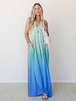 A flowy ombre maxi dress transitions from soft mint at the top to vivid cobalt blue at the hem, styled with layered turquoise jewelry, a macrame fringe bag, and flat sandals for an effortlessly free-spirited warm-weather look.