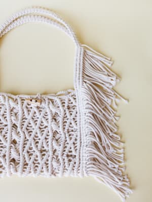 A cream macramé tote bag with knotted rope detailing, fringe trim along the side, and a small wooden toggle closure — photographed flat against a soft yellow backdrop with the kind of quiet handmade energy that feels both artsy and everyday.