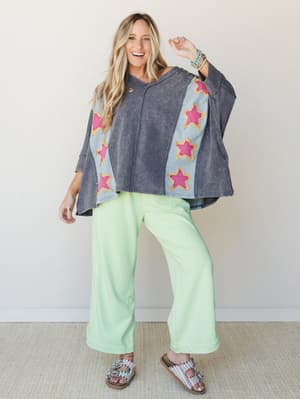 A smiling model in a oversized acid-wash poncho top with bold star appliqués paired with wide-leg mint green pants and embellished sandals — the whole look has this effortless, playful energy that feels like a weekend well spent.