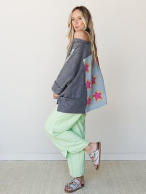 A model poses in profile wearing an oversized acid-wash sweatshirt with bold magenta star appliqués on a contrasting light blue back panel, paired with wide-leg mint green pants and embellished sandals — the whole look has a playful, free-spirited en