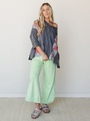 A relaxed full-body studio shot of a model in a washed navy oversized poncho top with colorblock patchwork and hot pink star appliqués, paired with mint green flare pants and embellished sandals — the whole look has that effortless, 'I just threw thi