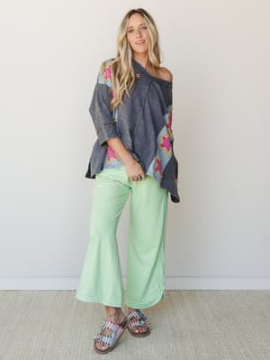 A relaxed full-body studio shot of a model in a washed navy oversized poncho top with colorblock patchwork and hot pink star appliqués, paired with mint green flare pants and embellished sandals — the whole look has that effortless, 'I just threw thi