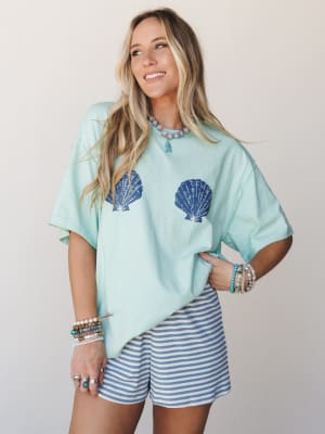 A sun-happy outfit pairing an oversized mint tee printed with navy scallop shells alongside blue-and-white striped shorts, styled with layers of beaded bracelets and a turquoise pendant necklace — the kind of look that feels equally at home on a boar