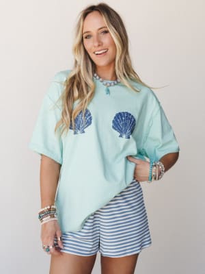 An oversized mint tee with navy seashell graphics is paired with blue-and-white striped shorts and layered beaded jewelry — casual, coastal, and the kind of outfit you'd pack first for a beach trip.