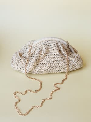 A cream woven raffia clutch with a gathered pouch silhouette and a gold chain strap laid casually against a soft warm-toned background — the kind of bag that makes a simple sundress feel intentional.