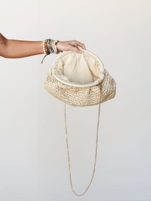A woven straw clutch held open by a bracelet-stacked hand against a clean light background — the gold chain strap and cream fabric lining visible, giving it that easy summer-to-evening energy.