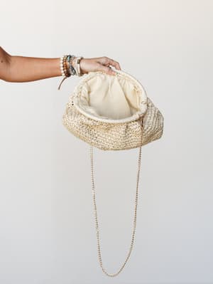 A woven straw clutch held open by a bracelet-stacked hand against a clean light background — the gold chain strap and cream fabric lining visible, giving it that easy summer-to-evening energy.