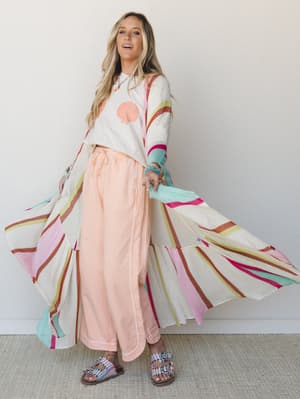 A model spins out the dramatic floor-length panels of a multicolor striped mesh duster, layered over a shell-graphic crop tee and soft peach wide-leg pants — the whole look has that easy, sun-soaked energy of someone who got dressed near the ocean.