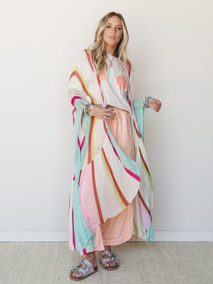 A bright, playful layered look built around a floor-length open-knit mesh duster in a multicolor stripe pattern, worn over a seashell graphic tee and a soft peach maxi skirt — the whole outfit has that easy, colorful energy of a beach town afternoon.