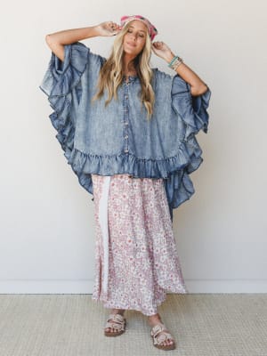 A breezy, full-body shot of a model in an oversized ruffled denim poncho layered over a flowy floral maxi skirt — the kind of outfit that looks like it happened effortlessly but definitely did not.