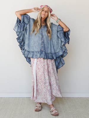A breezy, full-body shot of a model in an oversized ruffled denim poncho layered over a flowy floral maxi skirt — the kind of outfit that looks like it happened effortlessly but definitely did not.