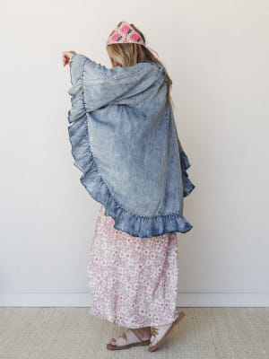 Shot from behind, a model fans out an oversized acid-wash denim jacket with dramatic ruffle trim, layered over a flowy pink floral maxi skirt — topped off with a crochet strawberry headband that ties the whole playful, boho-girl look together.