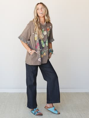 A laughing model pairs an oversized washed-brown butterfly graphic tee with wide-leg dark denim and sky blue sandals — the kind of effortless, personality-forward outfit that feels like a really good Saturday.