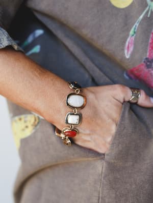 A close-up detail shot of a gold chain bracelet with mixed stone settings — cream, white marble, and rust red — styled against a taupe linen skirt and a floral print top, with a stacked ring just visible on the finger.