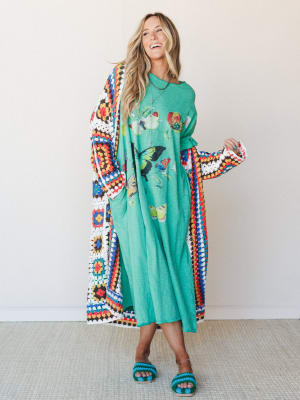 A woman laughing in a kelly green butterfly graphic maxi dress layered under a floor-length granny square crochet duster, styled with matching teal crochet slide sandals — the whole look has that 'yes, I dressed intentionally and I'm having a great t