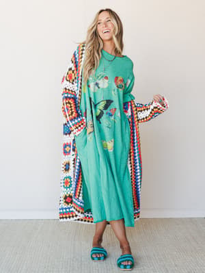 A woman laughing in a kelly green butterfly graphic maxi dress layered under a floor-length granny square crochet duster, styled with matching teal crochet slide sandals — the whole look has that 'yes, I dressed intentionally and I'm having a great t