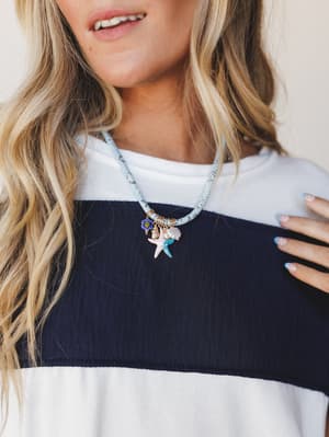 A close-up lifestyle shot of a charm necklace with starfish, seashell, and flower pendants on a light blue beaded cord, layered over a navy and white color-block ribbed top — easy, coastal, and just the right amount of playful.