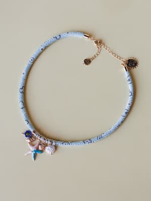 A fabric-wrapped cord choker in soft light blue with a delicate printed pattern, layered with ocean-inspired charms — a starfish, seashell, flower bead, and pearl — finished with rose gold hardware and a peace sign tag.