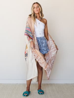 A sun-kissed model layers a flowy mixed-print kimono over a one-shoulder white lace crop top and periwinkle floral shorts, accessorized with stacked turquoise jewelry and crochet sandals — the whole look has that effortless 'grabbed everything off th