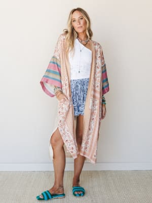A flowy floral kimono in soft blush and cream layers over a white lace crop top and blue floral shorts, styled with stacked beaded bracelets, a pendant necklace, and teal crochet slide sandals — the kind of outfit that looks effortless but absolutely