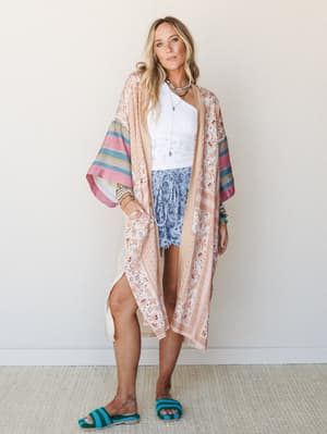 A flowy floral kimono in soft blush and cream layers over a white lace crop top and blue floral shorts, styled with stacked beaded bracelets, a pendant necklace, and teal crochet slide sandals — the kind of outfit that looks effortless but absolutely