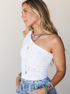 A side-profile shot of a model in a white one-shoulder lace crop top paired with a floral periwinkle midi skirt, layered with colorful beaded necklaces and stacked stone bracelets — the kind of outfit that feels equally at home at a summer concert or