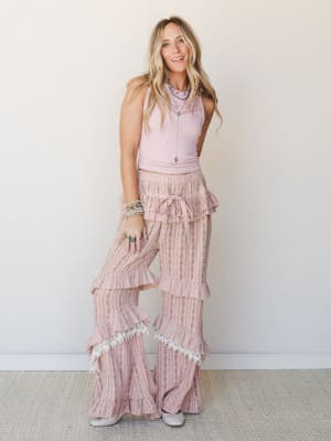 A blonde model wearing a ribbed blush crop tank paired with ultra-wide flare pants layered in ruffles, lace trim, and a floral stripe print — the whole look has a vintage-romantic, free-spirited energy styled with turquoise jewelry and silver flats.