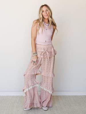 A blonde model wearing a ribbed blush crop tank paired with ultra-wide flare pants layered in ruffles, lace trim, and a floral stripe print — the whole look has a vintage-romantic, free-spirited energy styled with turquoise jewelry and silver flats.