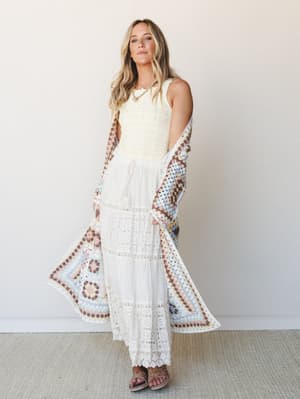A breezy full-length look layered with intention — a white eyelet and lace tiered maxi dress paired with a granny square crochet duster in soft pastels, styled with strappy sandals and a layered necklace against a clean studio wall.