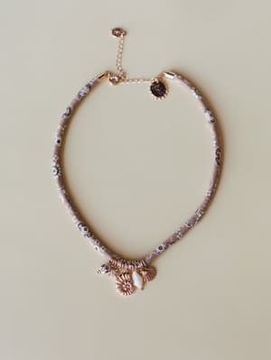 A fabric-wrapped charm choker in dusty mauve with a delicate floral print, finished with rose gold hardware and a cluster of charms — a sunburst, seashell, scallop shell, and speckled bead — laid flat on a soft sage-cream background.