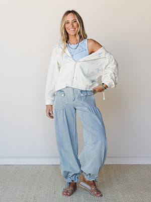 A sun-kissed blonde model layers a cream butterfly-print zip jacket over a light blue ribbed tank, paired with oversized light-wash barrel-leg pants and clear studded sandals — the whole look feels like a breezy Saturday with nowhere to rush.
