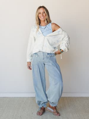 A sun-kissed blonde model layers a cream butterfly-print zip jacket over a light blue ribbed tank, paired with oversized light-wash barrel-leg pants and clear studded sandals — the whole look feels like a breezy Saturday with nowhere to rush.