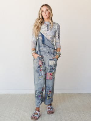 A layered boho two-piece moment — a floral print mesh long-sleeve top tucked under a wide-leg patchwork denim overall, covered in embroidered flowers and mixed fabric patches, styled with beaded bracelets and embellished sandals against a clean studi
