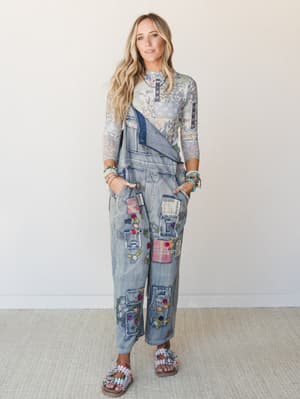 A layered boho two-piece moment — a floral print mesh long-sleeve top tucked under a wide-leg patchwork denim overall, covered in embroidered flowers and mixed fabric patches, styled with beaded bracelets and embellished sandals against a clean studi