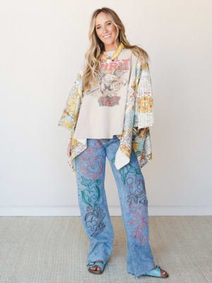 A full-body model shot of a layered boho outfit — graphic eagle tee, a patchwork-print open kimono with wide sleeves, and wide-leg embroidered jeans with colorful swirling designs, finished with turquoise sandals against a clean studio wall.