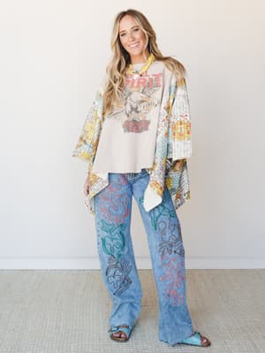 A full-body model shot of a layered boho outfit — graphic eagle tee, a patchwork-print open kimono with wide sleeves, and wide-leg embroidered jeans with colorful swirling designs, finished with turquoise sandals against a clean studio wall.
