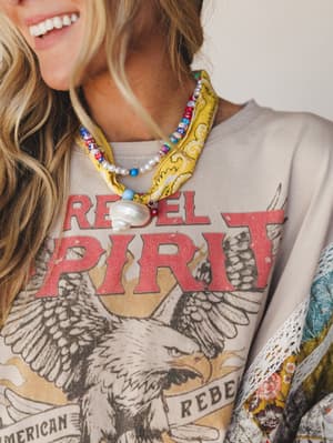 A close-up of a layered boho look built around a sand-toned graphic sweatshirt with a distressed eagle and 'Rebel Spirit' print, styled with a yellow paisley bandana scarf, a pearl and mixed bead necklace with a shell pendant, and a glimpse of a flor