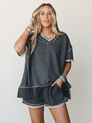 A relaxed charcoal matching set — oversized v-neck top and mini shorts with exposed contrast stitching — styled with stacked turquoise bracelets and colorful beaded necklaces for that effortless, personality-forward kind of cozy.