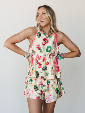 A laughing blonde model rocks a cream novelty-print matching set — halter top and ruffled shorts covered in tomatoes, hot sauce bottles, and garden motifs — styled with stacked beaded bracelets and a colorful beaded necklace against a clean studio ba