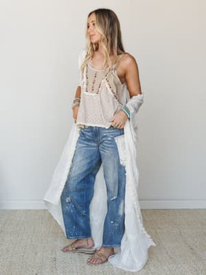 A breezy, layered look — embroidered cami, flowy white duster, and lived-in wide-leg jeans styled with stacked turquoise bracelets and slide sandals, shot against a clean indoor wall with that effortless 'just threw this on and it worked' energy.