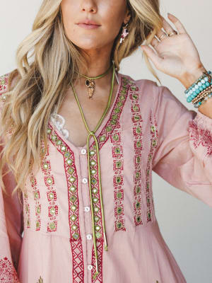 A close-up detail shot of a blush pink embroidered top with mirror accents and red geometric stitching, styled with a green lariat necklace, stacked bracelets, and layered rings — boho accessories doing all the right things.