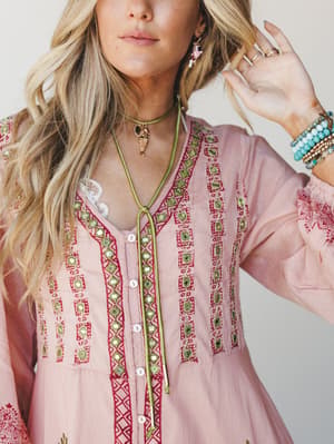 A close-up detail shot of a blush pink embroidered top with mirror accents and red geometric stitching, styled with a green lariat necklace, stacked bracelets, and layered rings — boho accessories doing all the right things.