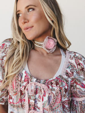 A dreamy close-up of a model in a paisley print top with a ribbon tie neckline, layered with a crochet choker necklace adorned with a soft pink organza flower — the whole look has that romantic, garden-party-meets-boho energy.