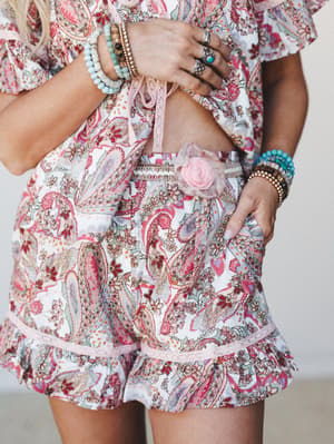 A close-up torso shot of a paisley print two-piece set — cropped top and ruffle-hem shorts — styled with stacked beaded bracelets, silver rings, and a fabric rose detail at the waistband, all feeling very collected and effortlessly boho.