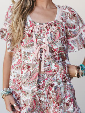 A close-up torso shot of a paisley-printed matching set — cropped tie-front top and skirt — layered with a coordinating open top, all in soft white and berry pink tones, accessorized with stacked beaded and turquoise bracelets.