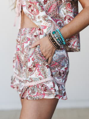 A close-up torso shot of a paisley print two-piece set — cropped top with tie details and ruffled shorts with lace trim — styled with a stacked beaded bracelet arm and hand tucked into a side pocket.