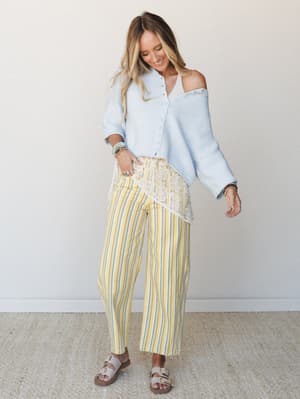 A sun-washed three-piece layered look — pale blue cropped cardigan worn off one shoulder over a white lace top, paired with wide-leg yellow stripe pants and buckle sandals, styled with stacked bracelets and an easy smile.
