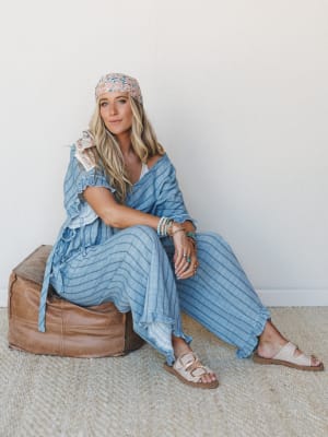 A relaxed, sun-warmed look — model sits on a leather pouf in a coordinated blue stripe set with ruffle trim, layered with a floral headscarf and stacked bracelets, the whole thing feeling like a slow afternoon somewhere coastal.