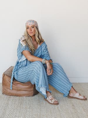 A relaxed, sun-warmed look — model sits on a leather pouf in a coordinated blue stripe set with ruffle trim, layered with a floral headscarf and stacked bracelets, the whole thing feeling like a slow afternoon somewhere coastal.
